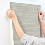 A person wearing a striped shirt is sticking a sheet of gray, horizontally textured Fountain Grass Camel Wallpaper Brown (60 Sq.Ft.) by York Wallcoverings onto a white wall. Only the person's arms and hands are visible as they smooth out the wallpaper, showcasing its easy installation and natural aesthetic.