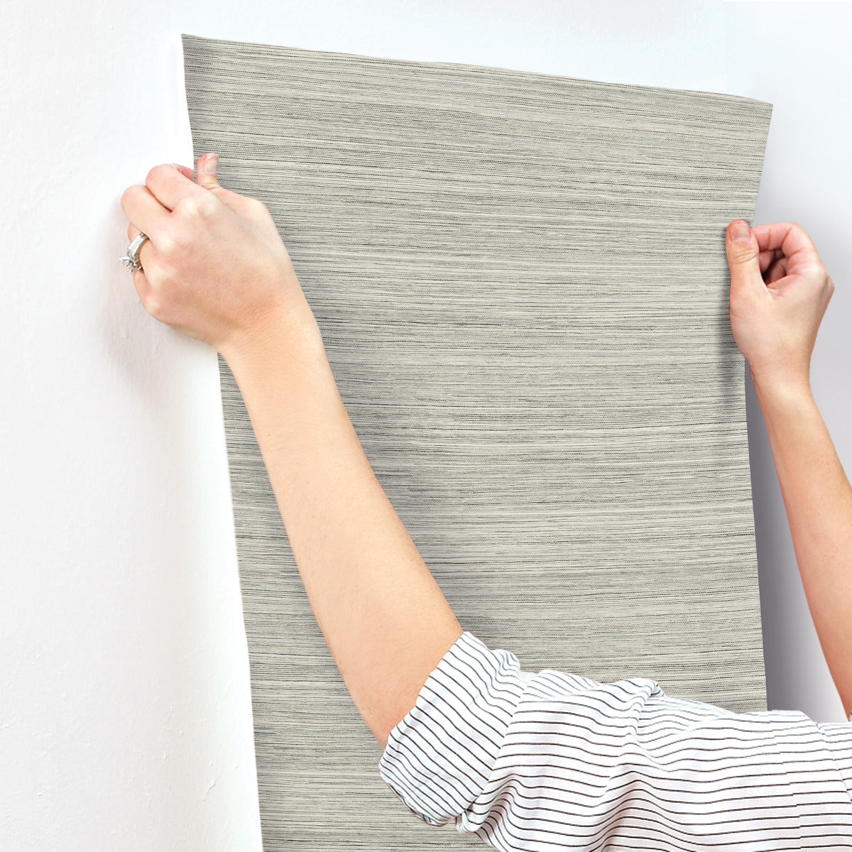 A person wearing a striped shirt is sticking a sheet of gray, horizontally textured Fountain Grass Camel Wallpaper Brown (60 Sq.Ft.) by York Wallcoverings onto a white wall. Only the person's arms and hands are visible as they smooth out the wallpaper, showcasing its easy installation and natural aesthetic.