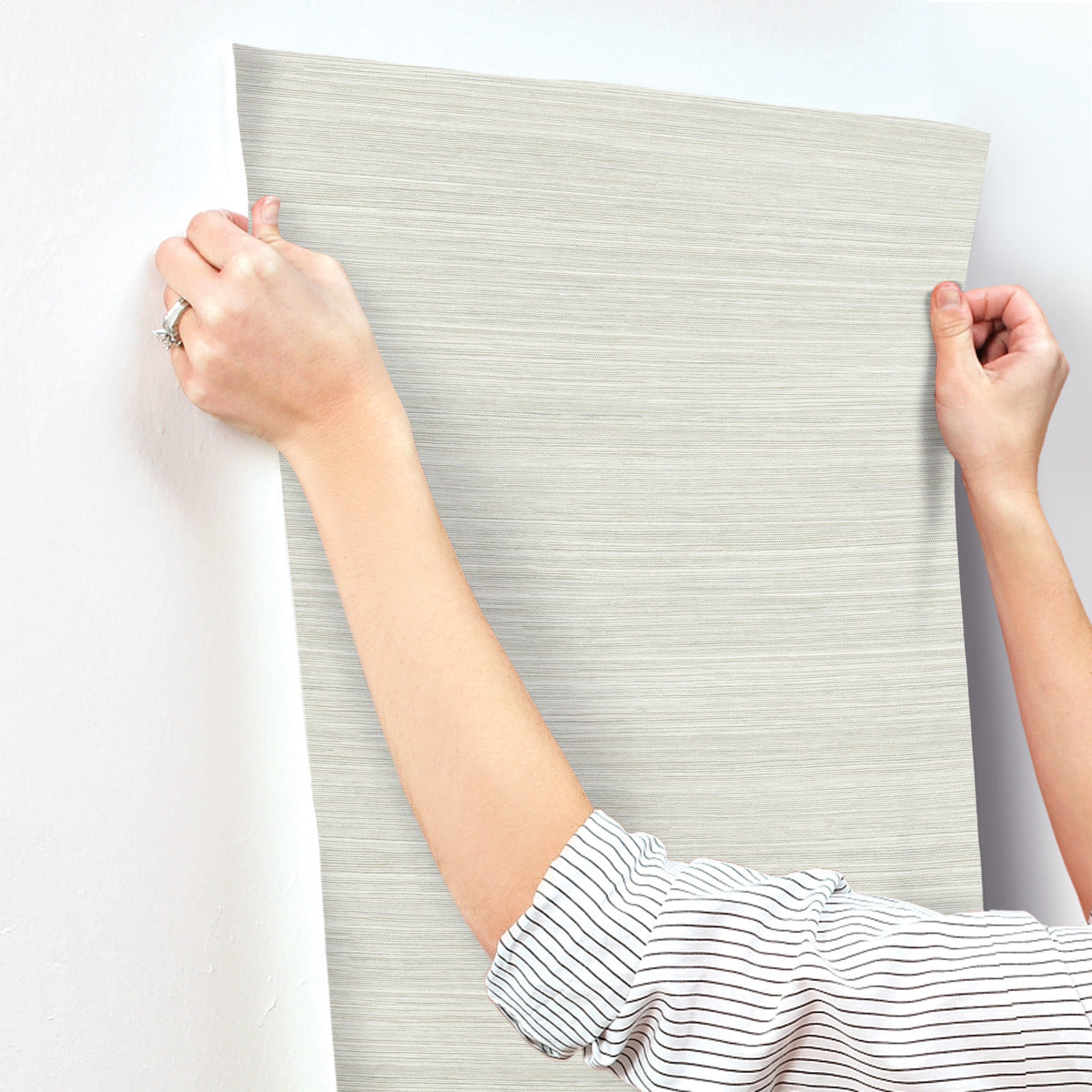 A person in a striped long-sleeved shirt is installing a piece of light grey wallpaper on a white wall. The person's hands are holding the top corners of the Fountain Grass Camel Wallpaper Brown (60 Sq.Ft.) by York Wallcoverings, ensuring it is being placed evenly. A ring is visible on one of their fingers, adding to the natural aesthetic.