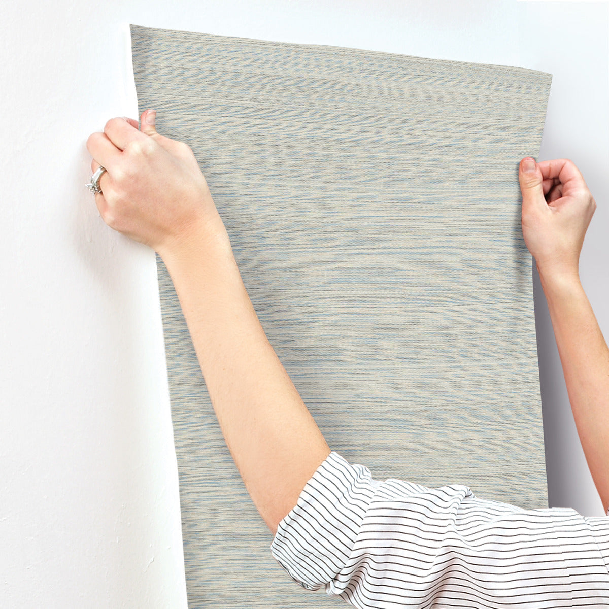 A person with a striped shirt is applying light grey, textured Fountain Grass Camel Wallpaper Brown (60 Sq.Ft.) by York Wallcoverings to a white wall. Their hands are smoothing out the wallpaper, showcasing its subtle horizontal pattern and natural aesthetic. A silver ring gleams on one finger, reflecting the easy installation process.