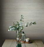 A minimalistic scene features a clear vase with eucalyptus branches on a light wooden table. Beside the vase are a small jar with a lid and a white bowl with some contents. The textured York Wallcoverings Fountain Grass Camel Wallpaper Brown (60 Sq.Ft.) serves as the backdrop, enhancing the serene, natural aesthetic.