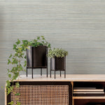 Two black metal planters on stands sit on a light-colored wood and wicker console. The left planter, with its trailing ivy plant, exudes a natural aesthetic, while the smaller planter on the right holds a succulent. Books are seen on the lower shelf of the console against a backdrop of Fountain Grass Smokey Blue Wallpaper Blue (60 Sq.Ft.) by York Wallcoverings.