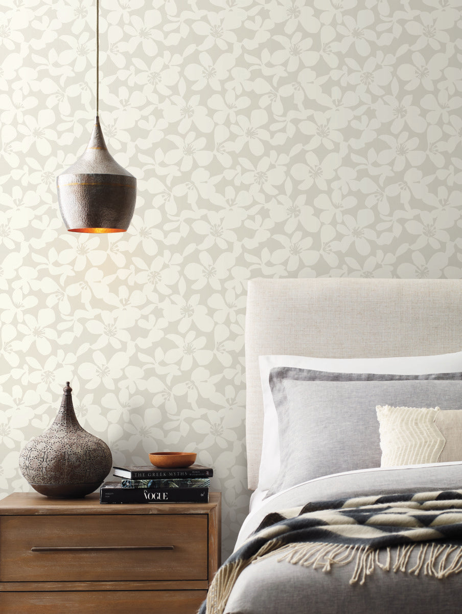 A cozy bedroom scene features a gray upholstered bed with gray and white bedding. A wooden nightstand holds a decorative vase, lit warmly by a hanging lamp above. The wall is adorned with York Wallcoverings' Free Spirit Wallpaper in Pearl, offering a subtle floral pattern and easy installation over 60 square feet of space.