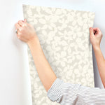 A person with rolled-up sleeves is applying York Wallcoverings' Free Spirit Wallpaper Pearl, which features a durable floral pattern of light beige flowers, to a white wall. This wallpaper covers 60 square feet and offers easy installation, making it perfect for refreshing any room.