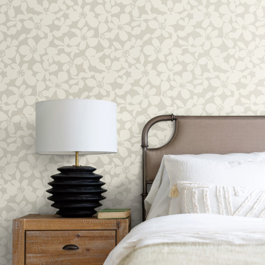 A bedroom scene features a black, textured lamp on a wooden nightstand beside a bed. The bed is enhanced by a beige headboard and white bedding with tasseled cushions. The wall is elegantly styled with the durable Free Spirit Wallpaper Pearl by York Wallcoverings, adding an elegant touch to the serene space.