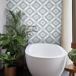 A modern bathroom showcases botanical elegance with a white oval bathtub in front of a **York Wallcoverings Garden Trellis Rain Wallpaper Blue, Grey (60 Sq.Ft.)**. Green potted plants are placed around the tub, enhancing the serene vibe. A wooden chair sits to the right, and beige curtains are partly drawn. The scene exudes tranquility and charm.