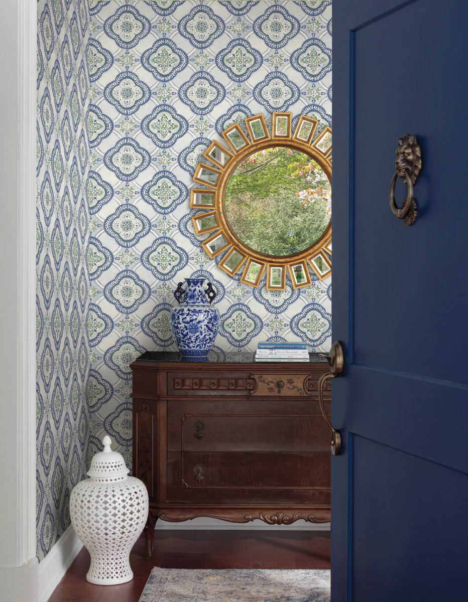 A small, stylish entryway features Garden Trellis Rain Wallpaper Blue, Grey (60 Sq.Ft.) by York Wallcoverings with a botanical elegance, a round golden mirror, a wooden dresser with ornate designs, a blue-and-white ceramic vase, and a white ceramic stool. A partially open navy blue door with a brass knocker reveals the inviting space.