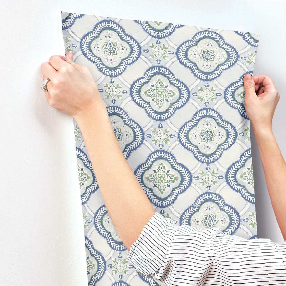 A person is applying the York Wallcoverings Garden Trellis Rain Wallpaper Blue, Grey (60 Sq.Ft.), adorned with intricate floral and geometric designs, to a white wall. They are using both hands to smooth out the wallpaper with ease. The person, wearing a white shirt with thin black stripes, exemplifies botanical elegance in their decorating process.