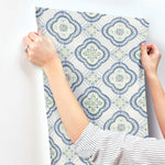 A person is applying the York Wallcoverings Garden Trellis Rain Wallpaper Blue, Grey (60 Sq.Ft.), adorned with intricate floral and geometric designs, to a white wall. They are using both hands to smooth out the wallpaper with ease. The person, wearing a white shirt with thin black stripes, exemplifies botanical elegance in their decorating process.