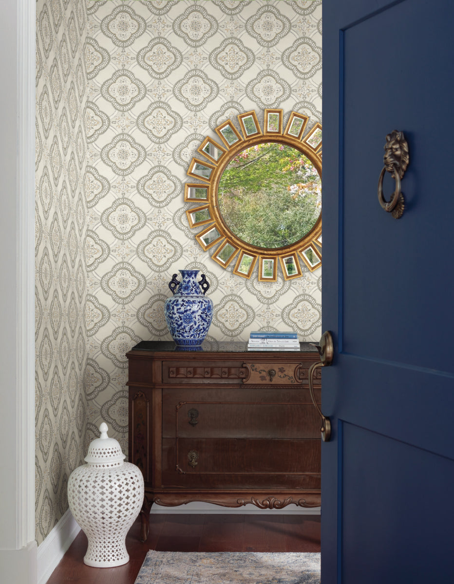 A stylish room features a dark wooden dresser against a York Wallcoverings' Garden Trellis Hazel Wallpaper Beige, Brown (60 Sq.Ft.) with intricate designs. Atop the dresser is a blue and white ceramic vase next to a round mirror with a gold frame. A white ceramic stool and a blue door with a brass knocker are in the foreground.