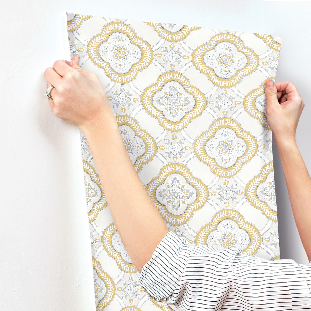 A person with rolled-up sleeves is installing York Wallcoverings Garden Trellis Hazel Wallpaper Beige, Brown (60 Sq.Ft.) with a gold and white ornate pattern. The removable wallpaper features decorative floral motifs inside oval shapes. The person's wrists and hands are visible as they smooth the wallpaper onto a white wall, adding botanical elegance to the space.