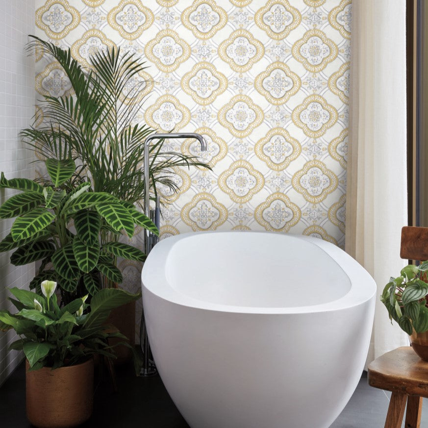 A freestanding white bathtub sits before a bright window with sheer curtains. The wall behind it features Garden Trellis Rain Wallpaper Blue, Grey (60 Sq.Ft.) by York Wallcoverings. Potted tropical plants, including a peace lily, are arranged around the bathtub, adding greenery and an easy touch of nature to the space.