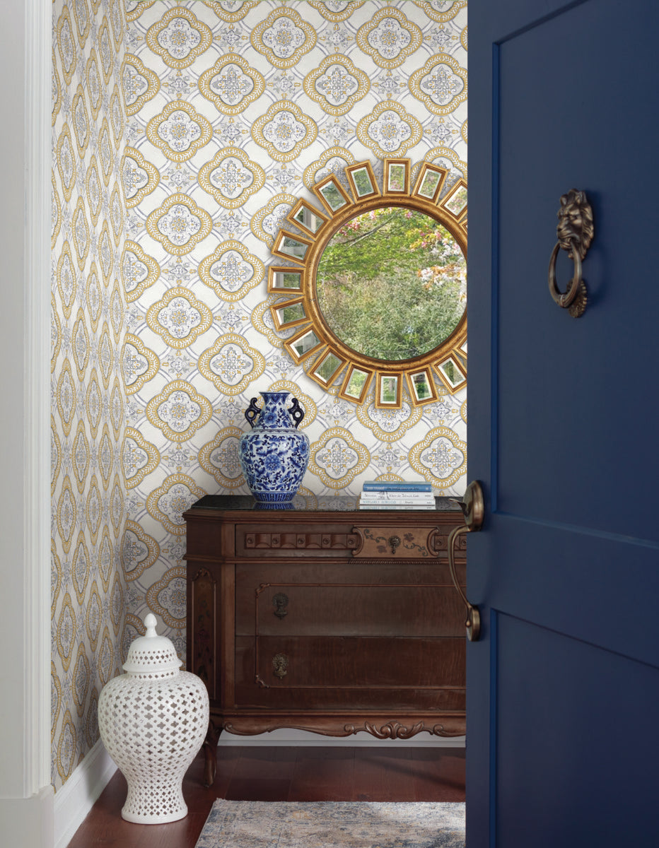 A corner of the room exudes botanical elegance with a wooden side table bearing an intricate design, a blue and white porcelain vase, and a circular mirror with a decorative frame reflecting greenery. The York Wallcoverings Garden Trellis Ochre Wallpaper Yellow (60 Sq.Ft.) boasts easy installation and removal. A ceramic stool sits beside a slightly open blue door with a brass knocker.