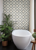 A modern bathroom features a white freestanding bathtub against a wall adorned with York Wallcoverings Garden Trellis Rain Wallpaper Blue, Grey (60 Sq.Ft.). The floor is dark, and to the left of the tub are potted green plants, adding botanical elegance. A wooden chair is placed on the right side, partially visible in the corner.