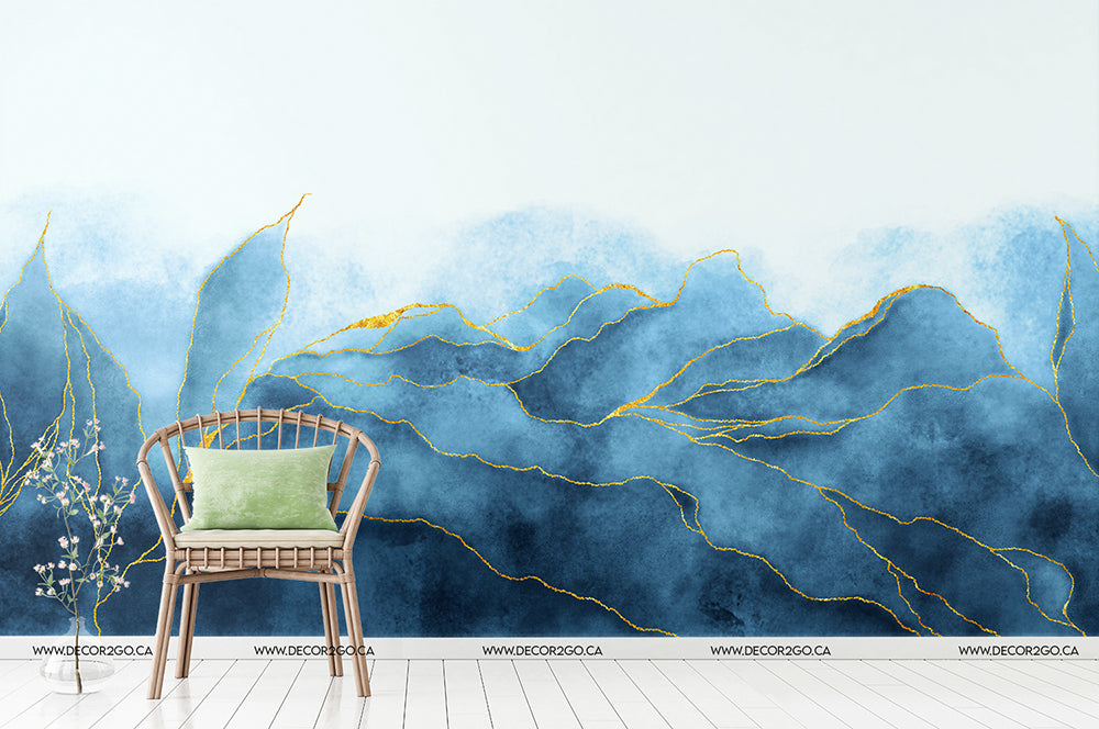 A modern interior features a wicker chair with a green cushion, placed on a white wooden floor. Behind the chair is Decor2Go Wallpaper Mural's Icy Glacier Wallpaper Mural in vivid detail, showcasing shades of blue with gold accents resembling mountain ranges or waves. A decorative plant sits beside the chair.