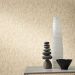 A minimalist scene features a tall white vase, a stack of smooth stones, and a black bowl on a light gray table against York Wallcoverings' Gold on Silver Shimmering Patina Wallpaper.