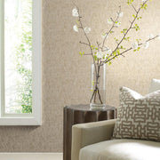 Living Room - A minimalist features a beige sofa, a patterned cushion, and a rustic wooden side table topped with a clear vase of blooming white branches. Sunlight filters through, softly illuminating York Wallcoverings' Gold on Silver Shimmering Patina Wallpaper with its metallic stipples