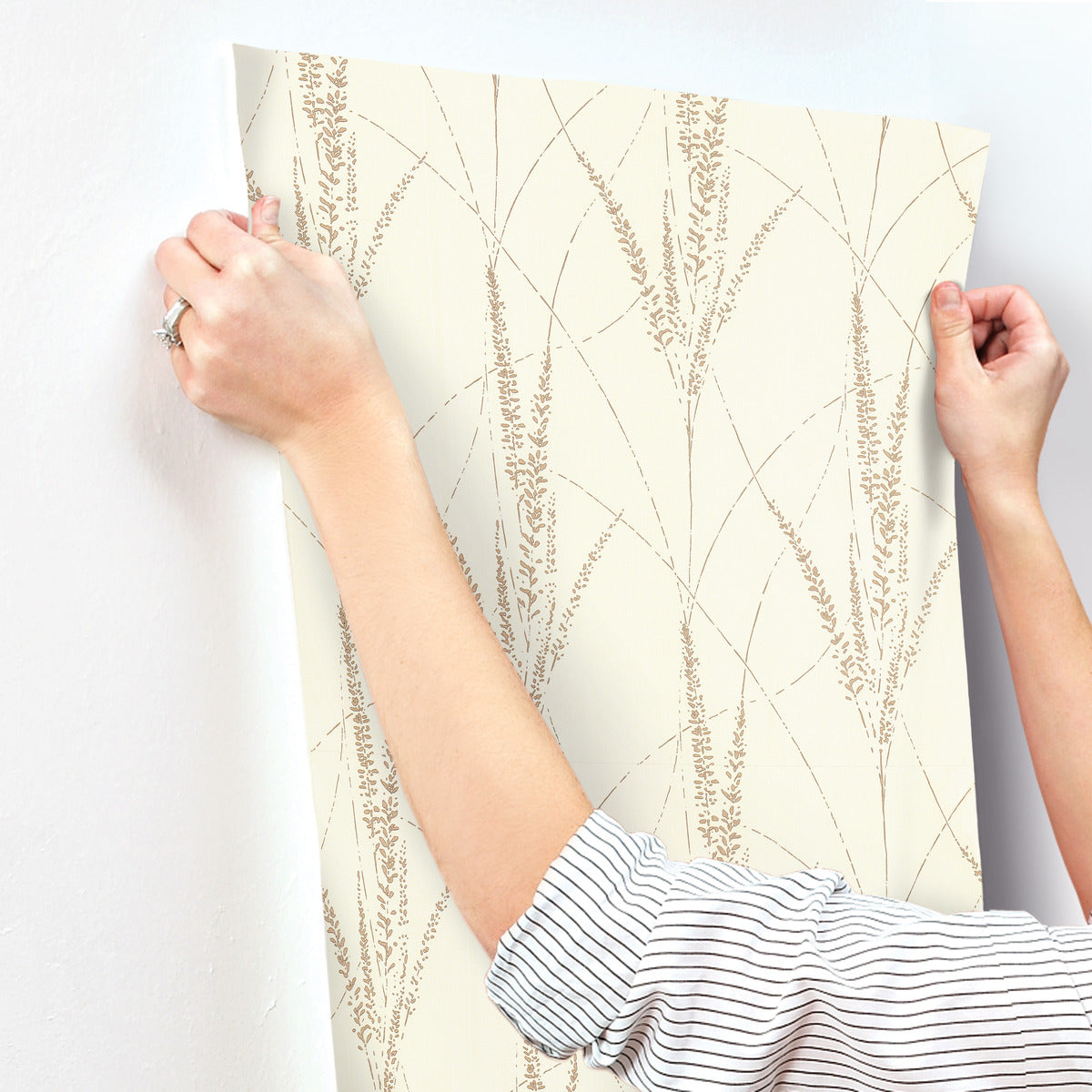 A person with rolled-up sleeves applies York Wallcoverings' Graceful Willow Cotton Wallpaper (60 SqFt) to a white wall, their hands smoothing the brown grass pattern with careful attention to detail.