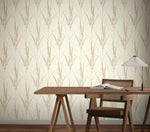 A wooden desk with an open notebook and lamp stands before York Wallcoverings' Graceful Willow Cotton Wallpaper, featuring a cream-colored botanical damask with a vertical wheat pattern. A wooden chair with a woven back is at the desk.