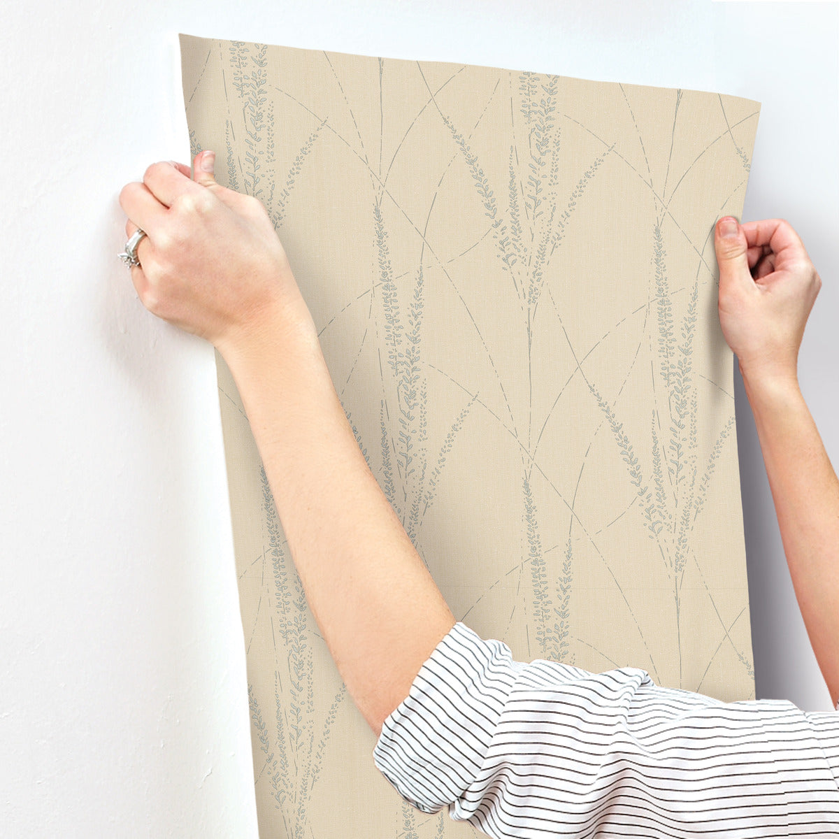A person with a striped sleeve applies York Wallcoverings' Graceful Willow Putty Wallpaper (60 SqFt), which features a subtle light gray botanical design, to a white wall. Only their hands and arms are visible.