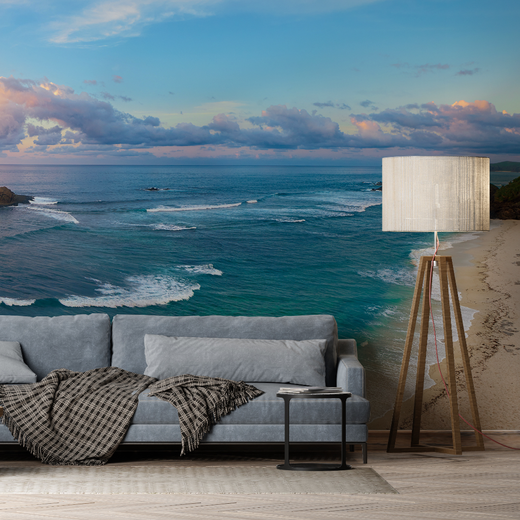 A cozy living room with a modern gray sofa and a stylish floor lamp, featuring a Decor2Go Wallpaper Mural that overlooks a stunning beach scene with rolling waves and a vibrant sunset.