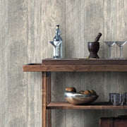 A wooden shelf against a York Wallcoverings Grey Rustic Wood Wallpaper (56 SqFt) wall displays a bottle of clear liquid, a filled martini glass, two empty martini glasses, a mortar and pestle, and a wooden bowl with apples. The shelf below holds a metal bowl with fruit and a stack of books.