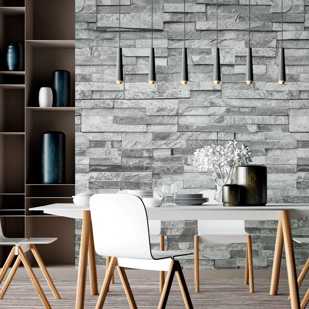 A modern dining area featuring a white table with wooden legs, complemented by white chairs. An elegant atmosphere is created by the Decor2Go Wallpaper Mural Grey Stone Wall Wallpaper Mural used as the backdrop, adorned with hanging pendant lights.