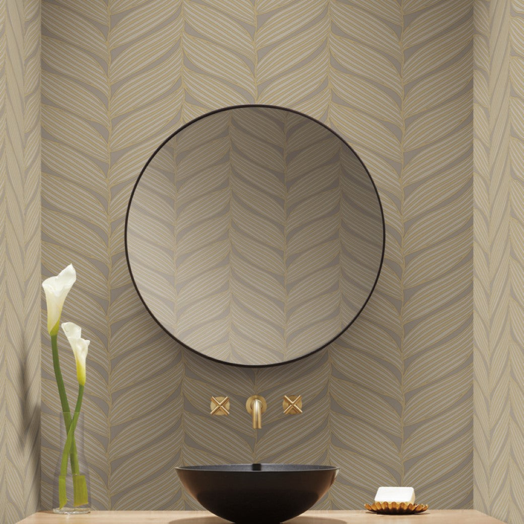 A modern bathroom features a round mirror, York Wallcoverings' Grey & Gold Luminous Leaves wallpaper, and a black vessel sink. Two gold faucets from the Modern Metals Collection adorn the wall. A minimalistic soap dish and vase of white flowers enhance the wooden countertop's elegance with metallic accents.