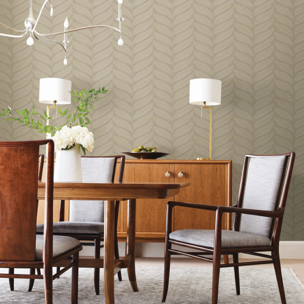 A dining room with a wooden table and four chairs, featuring a sideboard adorned with York Wallcoverings' Grey & Gold Luminous Leaves Wallpaper, while metallic accents in the modern chandelier complement the herringbone pattern wallpaper to add elegance.