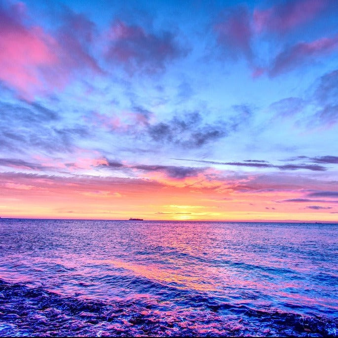 Vivid sunset over the ocean, featuring a dynamic sky with shades of pink, purple, and blue reflecting on the water's surface, ideal for a Decor2Go Wallpaper Mural with distant silhouetted ships.