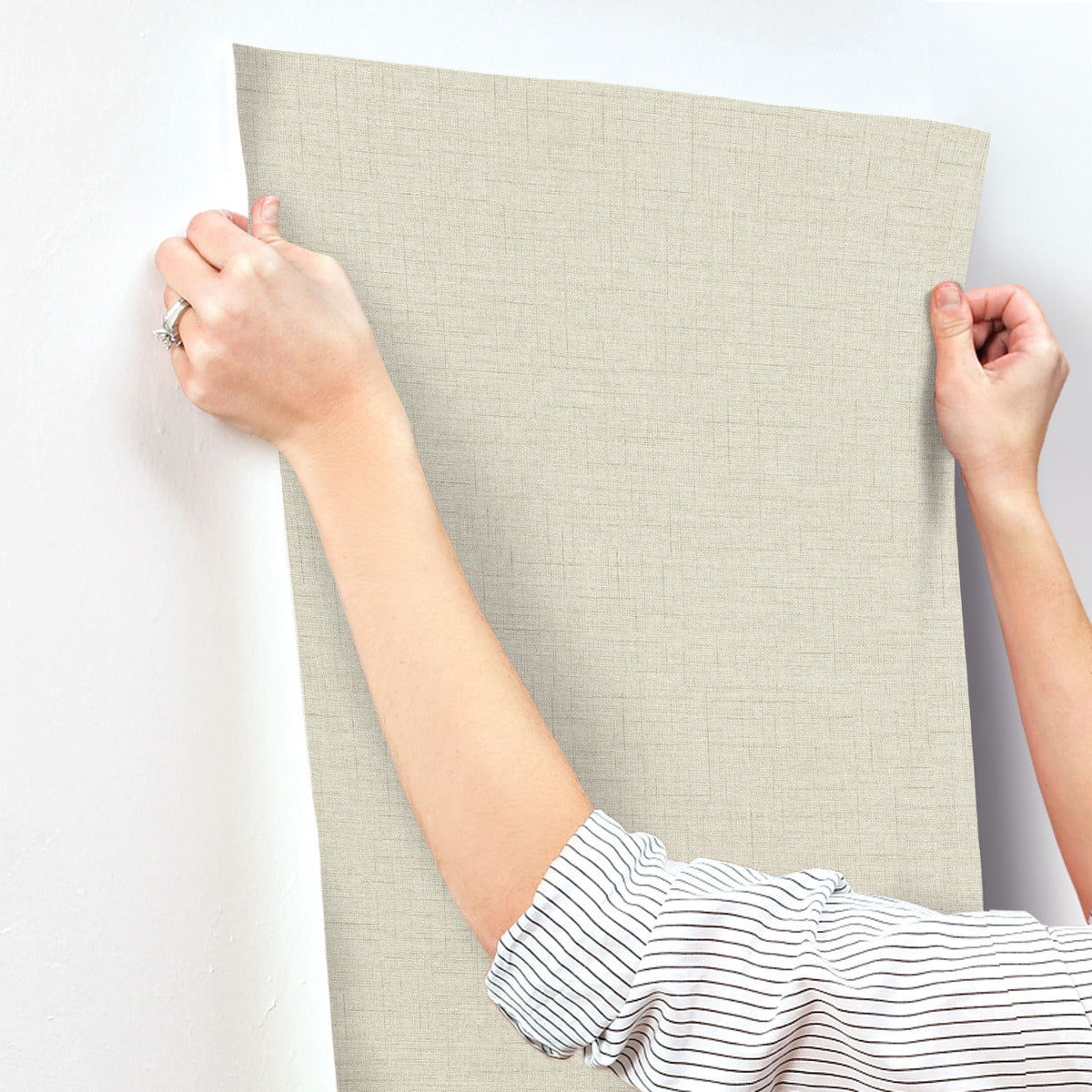 A person with rolled-up sleeves expertly applies York Wallcoverings' Hillswick Tweed Beige Wallpaper (60 SqFt) to a white wall, highlighting their hands and the careful wallpaper installation process.
