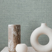Three minimalist vases—a tall marbled, a small round, and a large one with a circular hole—sit on light stone against York Wallcoverings' Hillswick Tweed Indigo Wallpaper (60 SqFt), featuring a soft blue-gray menswear fabric texture.