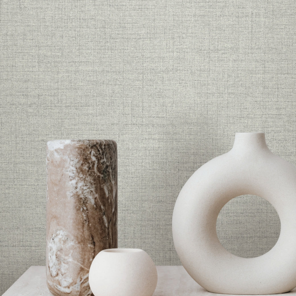 Three minimalist ceramic vases rest on a light surface, set against York Wallcoverings' Hillswick Tweed Pewter Wallpaper (60 SqFt); the vases include a round piece with a large hole, a marbled cylindrical vase, and a small spherical one.