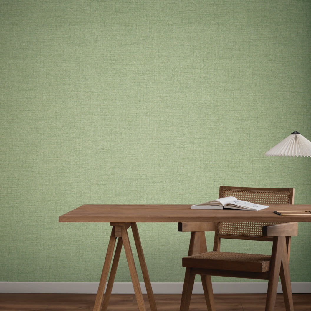A wooden desk with an open book and table lamp sits against a wall covered in Hillswick Tweed Shamrock Wallpaper by York Wallcoverings. A wooden chair with a woven back is at the desk, and the floor is brown wood.