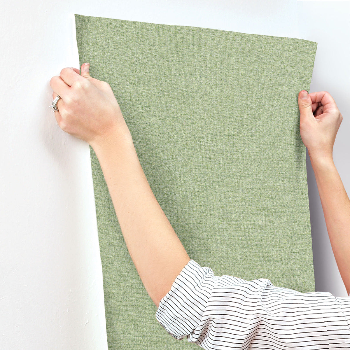 A person in a striped shirt is applying York Wallcoverings' Hillswick Tweed Shamrock Wallpaper (60 SqFt), featuring a textured green pattern, to a white wall, smoothing it with their hands.