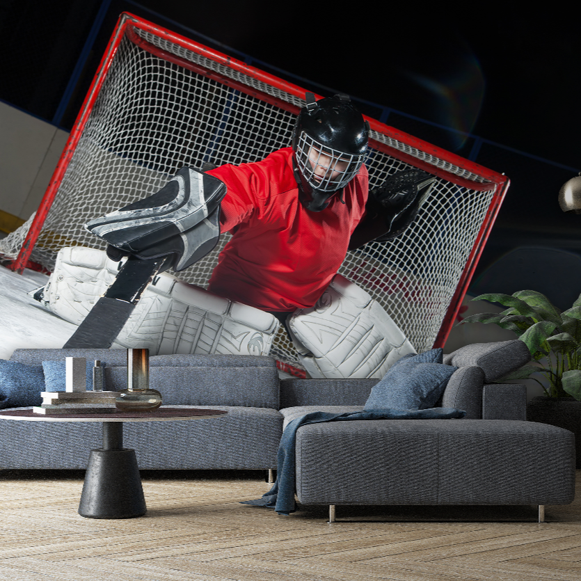 A Decor2Go Wallpaper Mural hockey goalie, in full gear and a red jersey, is crouched in a defensive position in front of a net that is set up in a living room with couches and a coffee table.