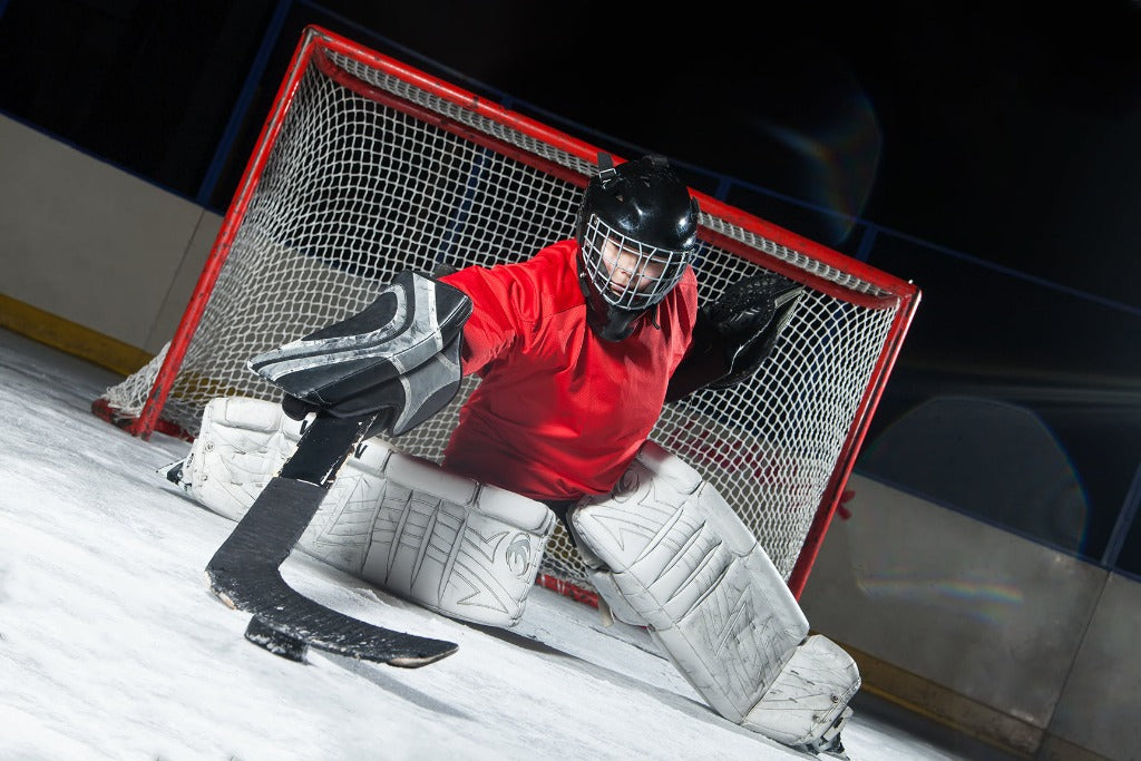 A Decor2Go Wallpaper Mural of a hockey goalie in red uniform and protective gear crouches in front of the goal on an ice rink, ready to block a shot under bright lights, captured in a high-quality image.