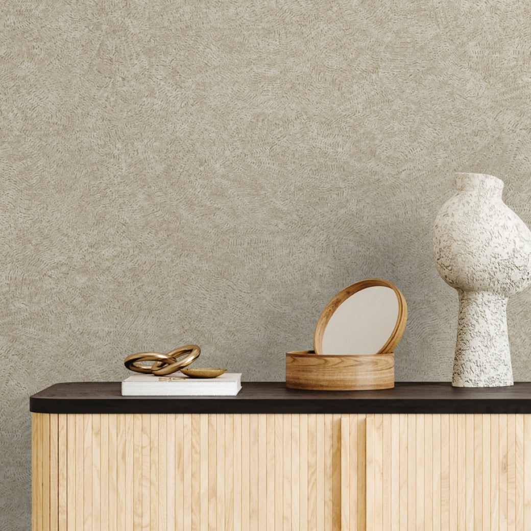 A minimalist sideboard with light wood doors displays a sculptural white vase, gold knot, and closed book against York Wallcoverings’ Impression Sand Wallpaper (60 SqFt), adding modern ripple texture ideal for contemporary interiors.