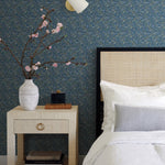 A bedroom showcases a beige nightstand with a white vase of pink flowers, books, and a clock. White and gray pillows adorn the bed. The Indigo Tapestry Wallpaper by York enhances the botanical design, complemented by a light fixture above.