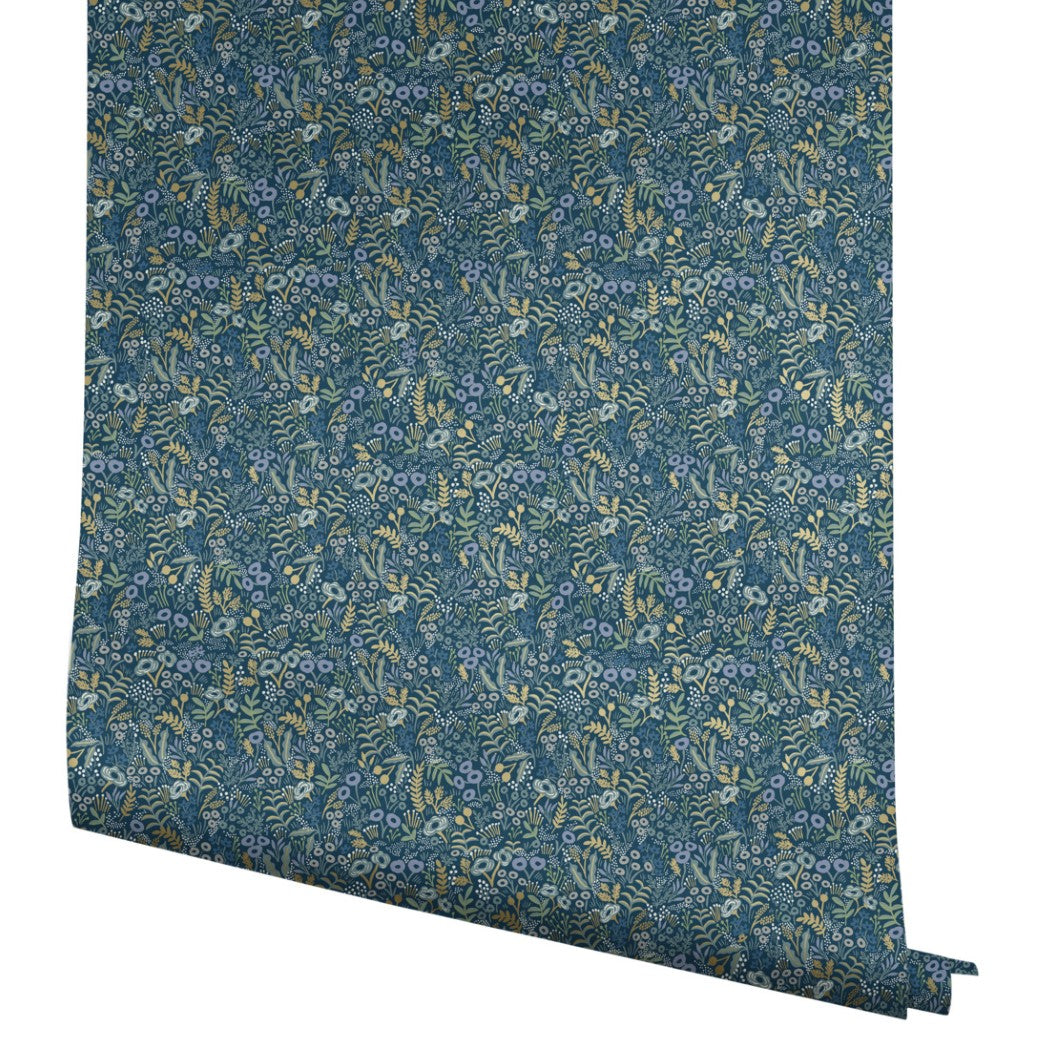 The Indigo Tapestry Wallpaper by York unrolls, displaying a complex botanical pattern of small blue flowers and green leaves on a dark blue backdrop, capturing every floral detail in the partially unfurled 60 SqFt roll.