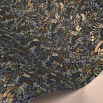 The Indigo Tapestry Wallpaper by York (60 SqFt) displays a dark blue background adorned with intricate botanical designs, featuring beige, green, and light blue leaves and flowers. Its partially curled form elegantly reveals the captivating floral artistry.