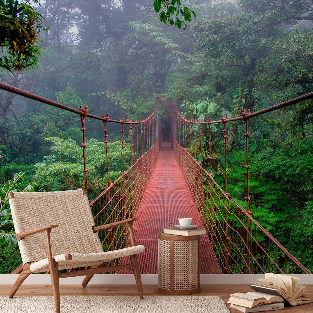 A cozy outdoor reading nook with a modern chair and side table, overlooking a long, narrow suspension bridge leading into a foggy, leafy environment. An open book rests on the ground featuring the Into the Forest Wallpaper Mural from Decor2Go Wallpaper Mural.
