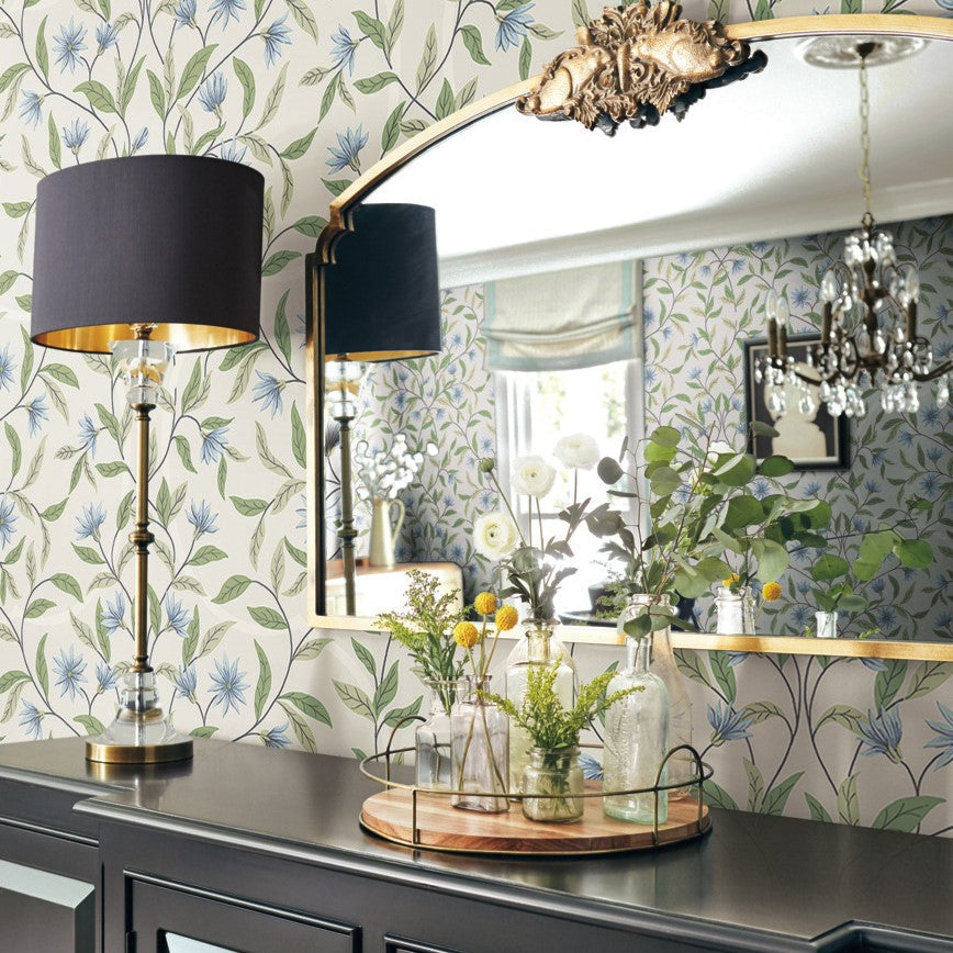 A room with York Wallcoverings Jasmine Cornflower Wallpaper Beige, Blue (60 Sq.Ft.) exudes botanical elegance. A large decorative mirror hangs above a dark wooden cabinet, holding a tray with assorted vases of yellow and white flowers. On the left, a tall table lamp with a black shade and gold accents adds to the charm, easy installation and removal.