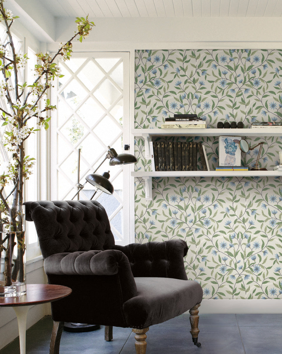 A cozy reading nook features a black velvet armchair beside a small wooden round table with a vase holding a blossoming branch. The white walls adorned with Jasmine Cornflower Wallpaper Beige, Blue (60 Sq.Ft.) by York Wallcoverings showcase floral patterns, and shelves hold books and decorative items. A window with a crisscross design allows natural light in.