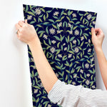 A person wearing a striped shirt is hanging a piece of Jasmine Indigo Wallpaper Blue (60 Sq.Ft.) with a navy blue background. The wallpaper, from the Botanical Elegance collection by York Wallcoverings, features green leaves and white flowers. Utilizing SureStrip technology, the person is pressing it against a white wall using both hands.