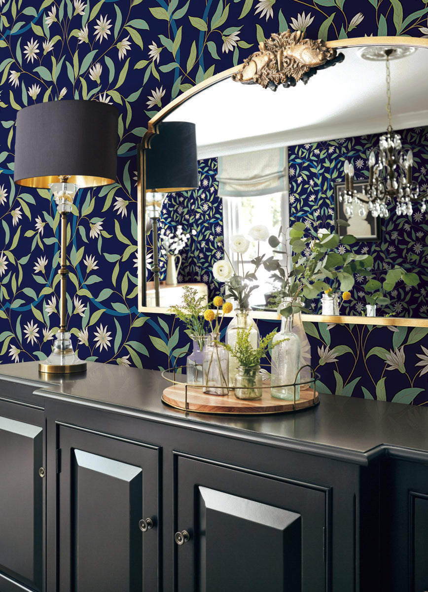 A dark-colored cabinet stands against a Jasmine Indigo Wallpaper Blue (60 Sq.Ft.) by York Wallcoverings with a large, ornate mirror above it. On the cabinet are decorative items, including glass vases with flowers and a tray. A tall, black and gold lamp with a shade adds a touch of Botanical Elegance.