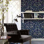 A cozy reading nook with a dark, tufted armchair beside a round, wooden side table. The background features a window with a diamond grid pattern and blue York Wallcoverings Jasmine Indigo Wallpaper Blue (60 Sq.Ft.). Two white shelves with books and decor items are mounted on the wall.