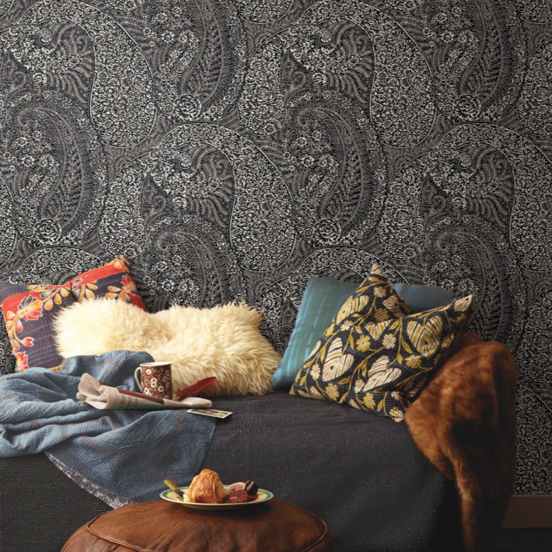 A cozy room corner showcases the Kashmir Dreams Paisley Wallpaper in black by York Wallcoverings. The couch is decorated with an assortment of patterned cushions, a fluffy white pillow, and a brown throw blanket. A wooden stool holds a mug and a plate of pastries, highlighted with subtle metallic accents.