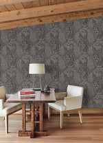 A cozy room featuring a wooden table, two cushioned chairs, and a table lamp is complemented by books and a small plant adorning the tabletop. The wall showcases the intricate Kashmir Dreams Paisley Wallpaper in black by York Wallcoverings, while wooden beams on the ceiling add subtle warmth to the space.