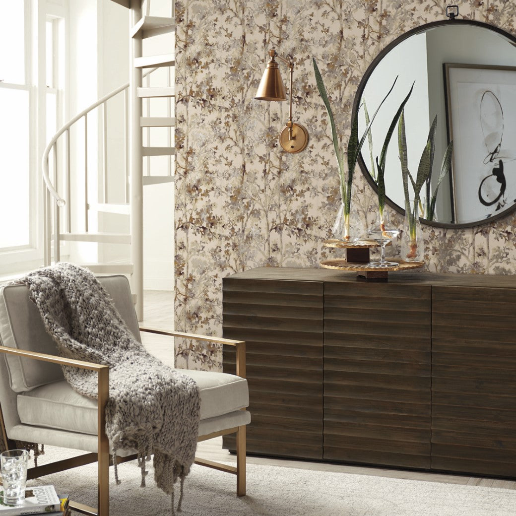 Living Room - A cozy features a beige armchair and knitted throw. On the right, a dark wooden dresser is topped with items from the Modern Metals Collection, including a copper vase. The Khaki & Multi Shimmering Foliage Wallpaper by York Wallcoverings hosts a round mirror, with a spiral staircase in view